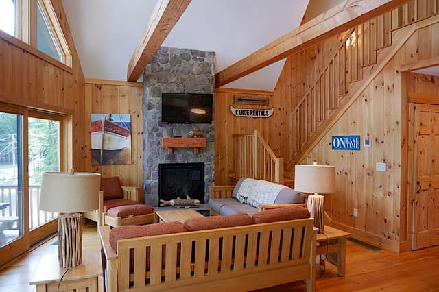 Bright, open-concept cabin living room. Highlights include knotty pine walls, a high beamed ceiling, a stone fireplace, and a wood staircase.