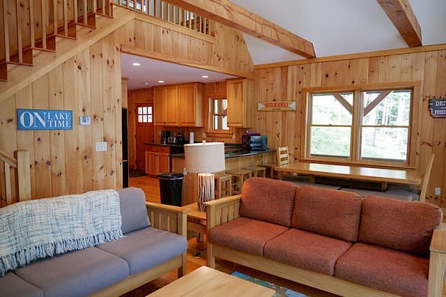 Cozy, open-concept cabin living space featuring knotty pine walls, wood-frame furniture, a dining area, and a loft staircase. The kitchen is visible in the back.