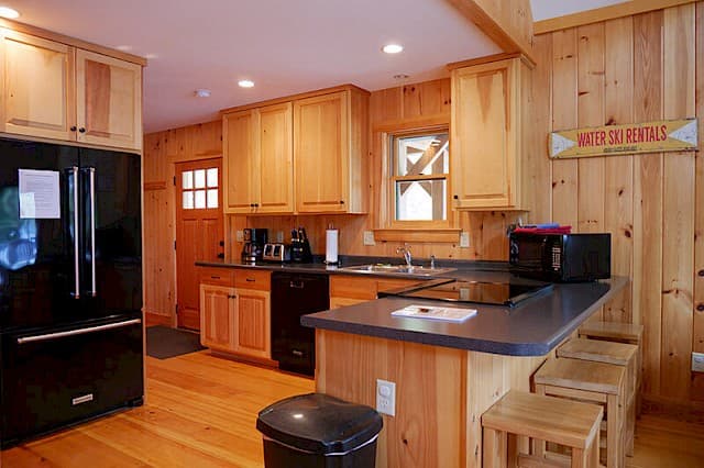 Fully equipped kitchen featuring natural pine walls and cabinets. Includes modern black appliances, a breakfast bar with seating, and a rustic wood floor.
