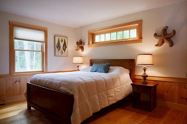 Cozy lake house master bedroom with a queen bed, wood flooring, pine trim, and a relaxing nautical theme. Features large and transom windows.
