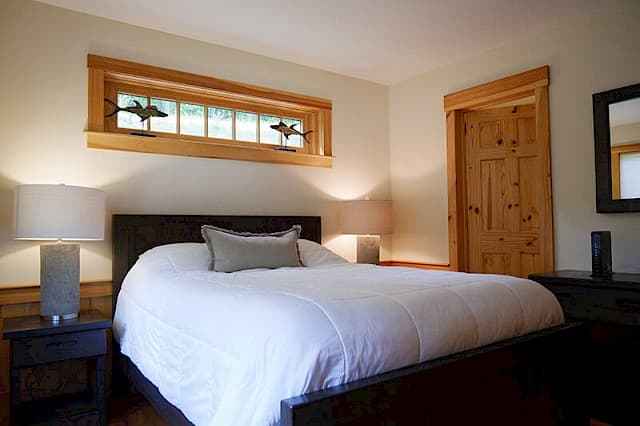 Serene master bedroom with a queen bed, modern lamps, and dark accents. Highlights include pine trim, a rustic pine door, and a unique leaded glass transom window.