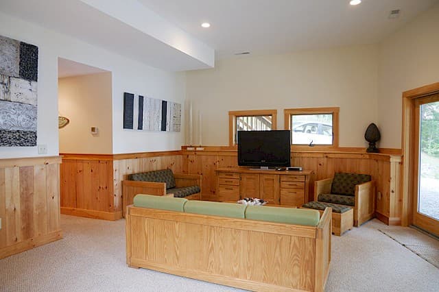 Spacious downstairs living room with a TV area, plush carpet, and light pine wainscoting. Features a couch, chairs, and a sliding glass door for outdoor access.