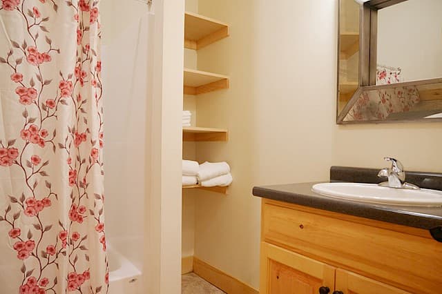 Bright, clean bathroom featuring a wood vanity, rustic mirror, and open storage shelves. A cheerful pink floral shower curtain adds a pop of color.