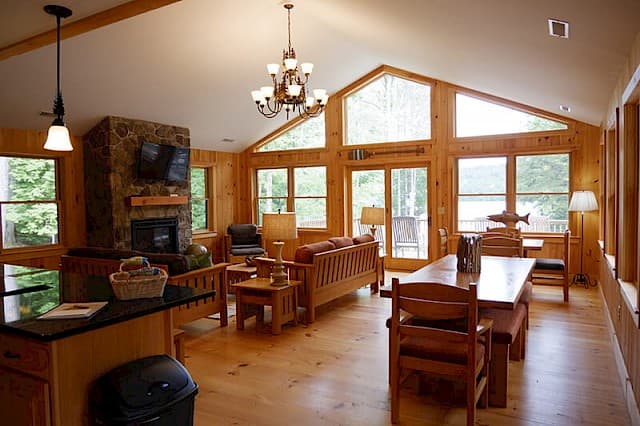 Stunning open-concept living space with a vaulted, beamed ceiling, knotty pine walls, and a large stone fireplace. Wall of windows offers bright lake views.