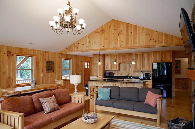 Warm, open-concept cabin interior featuring knotty pine paneling, a vaulted ceiling, two living room sofas, and a full kitchen with a breakfast bar.