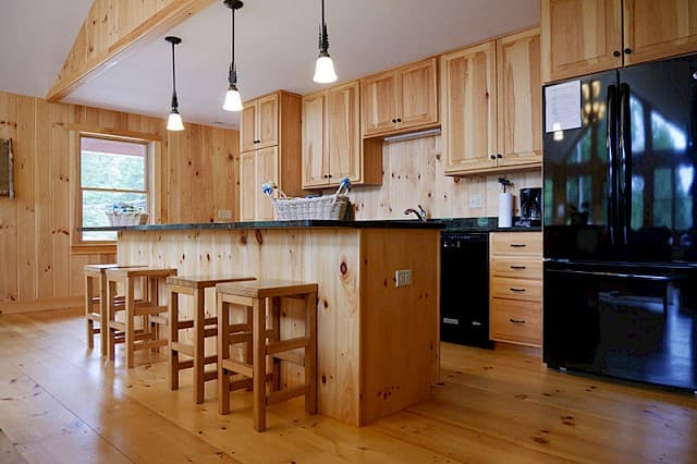 Spacious, modern cabin kitchen. Features knotty pine walls and cabinets, a large island with bar seating, and sleek black appliances and countertops.
