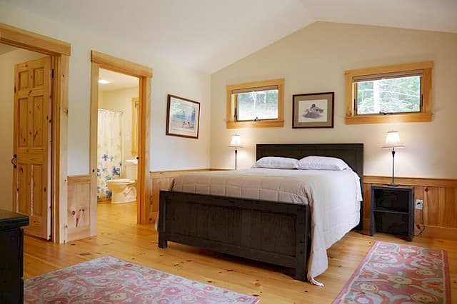 Spacious master bedroom with a vaulted ceiling, light wood floors, and pine wainscoting. Features a Queen bed and an ensuite full bath visible through the doorway.