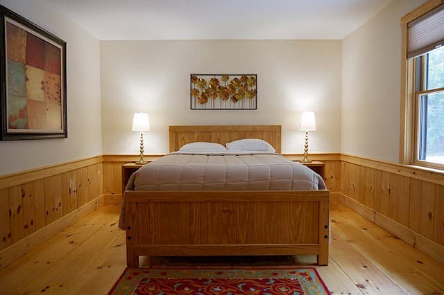 Relaxing queen bedroom featuring warm natural wood floors, bed frame, and pine wainscoting. Two matching lamps flank the bed.