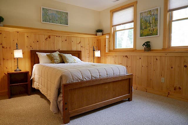 Warm and inviting queen bedroom. Features extensive knotty pine wainscoting, a wood bed frame, soft carpet, and twin reading lights.