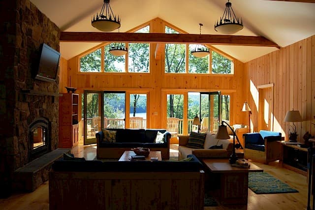 Stunning great room with a vaulted ceiling, exposed beams, and a massive stone fireplace. A wall of windows offers panoramic lake views from the sofa seating.