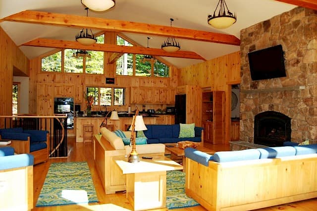Massive open-concept great room featuring a vaulted ceiling, exposed beams, and a huge stone fireplace. Knotty pine walls surround multiple seating areas.