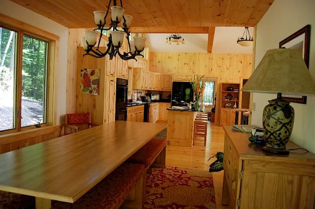 Open-concept dining and kitchen area. Features a very long pine dining table with benches, a chandelier, and extensive knotty pine paneling and cabinets.