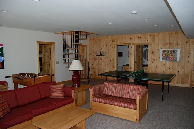 Large lower-level game room featuring a ping-pong table, foosball, and a comfortable seating area. Highlights include pine paneling and a spiral staircase.