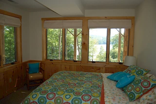 Bedroom with an incredible lake view through the wall of windows. Features bright, cheerful bedding, light pine wainscoting, and natural light.