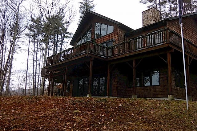 Grand, multi-story lake house exterior. Features dark wood siding, a stone chimney, expansive windows, and a wrap-around deck overlooking the wooded setting.