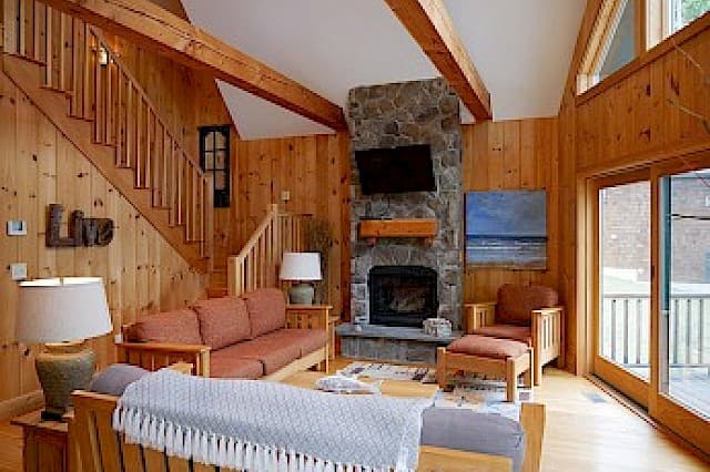 Cozy cabin living room featuring a dramatic stone fireplace, vaulted ceiling with beams, and knotty pine walls. A wood staircase leads up, and sliders open to a deck.