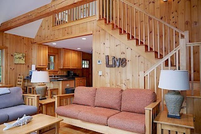 Cozy cabin living room with knotty pine walls and a vaulted ceiling. Features a prominent wooden staircase and an open doorway leading into the kitchen area.