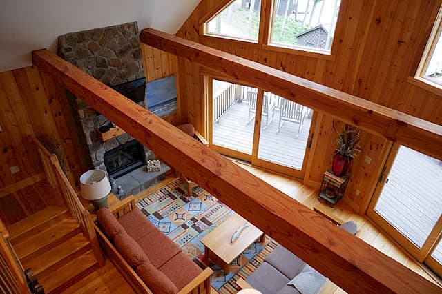 Unique loft view of the living room, showing the stone fireplace and vaulted ceiling. Exposed beams and pine walls frame the glass doors leading to the deck.