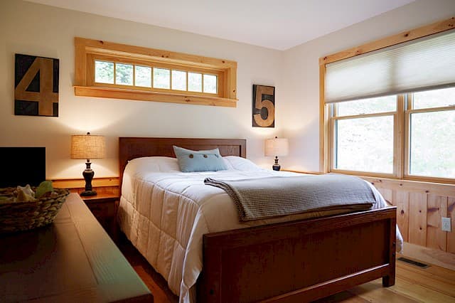 Bright, modern-rustic bedroom with a dark wood queen bed and dresser. Features a unique transom window, large side window, and light pine wainscoting/trim.