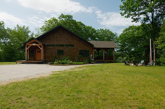 Exterior view of a large, rustic wood building with a prominent front entryway and a long, covered side porch. Surrounded by a green lawn and trees.