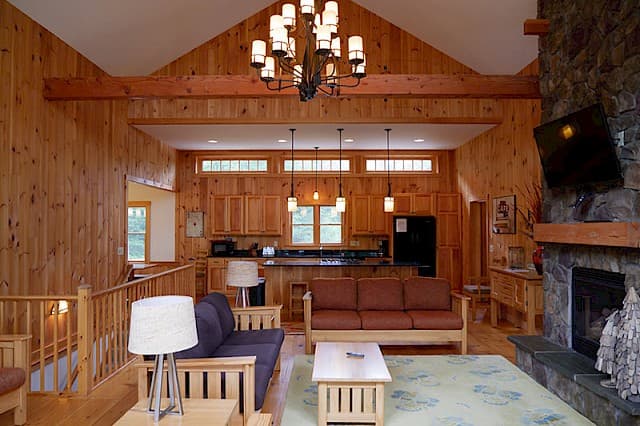 Bright, open-concept living area with a vaulted ceiling, cozy seating, and a large stone fireplace. The room features knotty pine walls and an integrated kitchen.