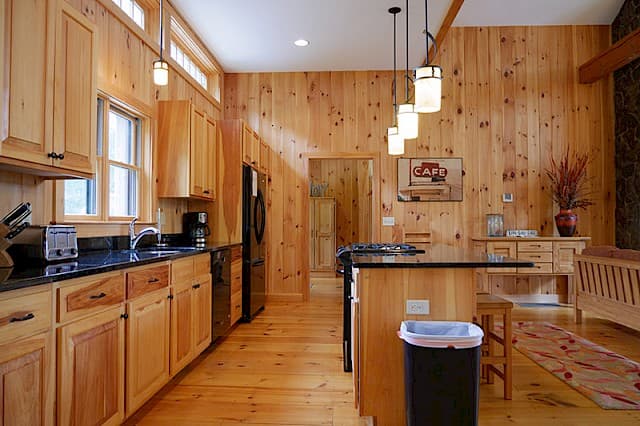Bright, modern galley kitchen with extensive knotty pine walls and cabinetry. Features black appliances, dark counters, and a central island with pendant lighting.