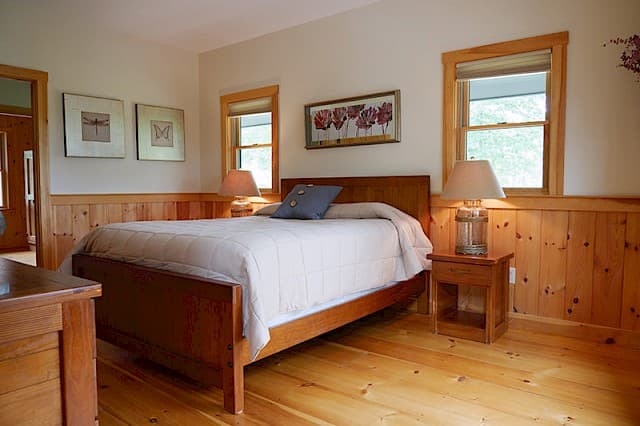Bright, welcoming queen bedroom featuring warm wood floors, a matching bed frame, and knotty pine wainscoting and window trim.