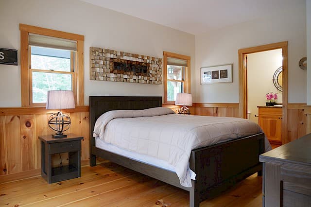 Inviting queen bedroom with light wood floors and rustic knotty pine wainscoting. Features a decorative mirror and an open doorway to the ensuite bath.