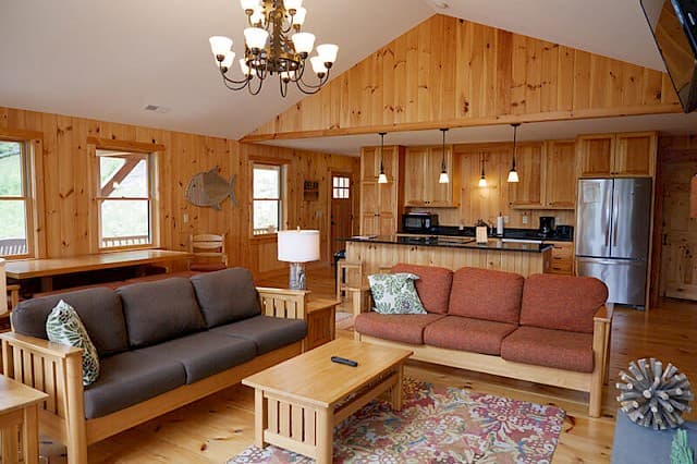 Warm, open-concept cabin living space. Features knotty pine walls, two sofas, a vaulted ceiling, and a full kitchen with a stainless steel fridge in the background.