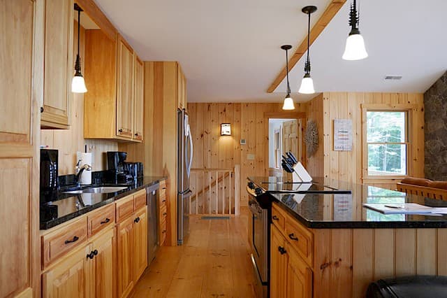 Bright kitchen with extensive pine paneling and modern black countertops. The island features pendant lights, and a staircase leads down in the background.