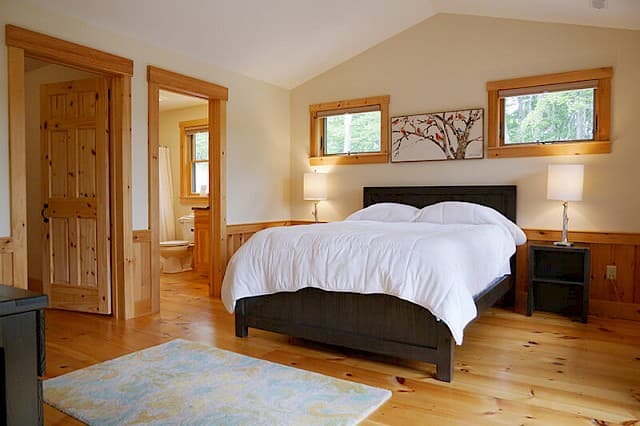 Spacious queen bedroom with a vaulted ceiling. Features light pine floors/wainscoting, a rustic door, and an open doorway to the private full bathroom.