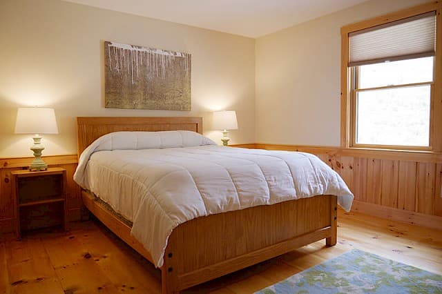 Bright, serene queen bedroom featuring a natural wood bed frame, rustic knotty pine wainscoting, and matching wood floors.