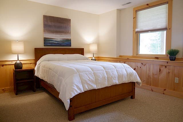 Tranquil queen bedroom featuring a wood bed frame and cozy carpet. Knotty pine wainscoting and window trim provide a warm, rustic cabin feel.