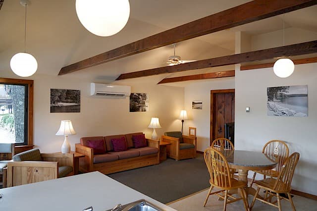 A cozy living space featuring exposed wooden beams, modern globe lights, a sofa, armchair, and a wicker dining set.
