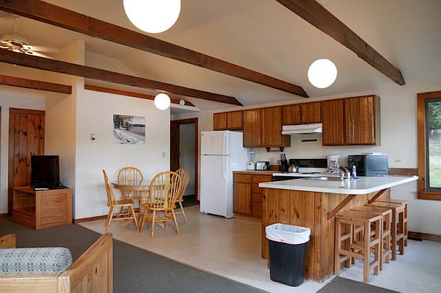 A well-lit kitchen and dining area with exposed wooden beams, modern globe lighting, wood cabinets, a white refrigerator, and a small breakfast bar.