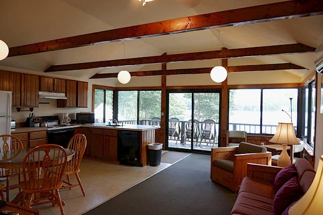 An open-plan cabin interior with exposed wooden beams, featuring a kitchen, dining area, and living space opening onto a deck with lake views.