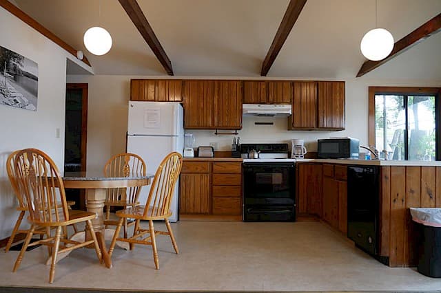 A cabin's kitchen and dining area with exposed wooden beams, featuring light wood cabinets, a white fridge, black stove, and a round wooden table.