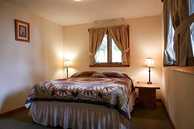 A cozy bedroom with a double bed covered by a patterned quilt, framed by two matching lamps on wooden nightstands and a small window.