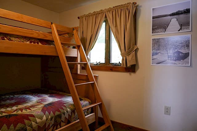 A simple cabin bedroom featuring natural wood bunk beds with a ladder, a small window with beige curtains, and two black and white prints.