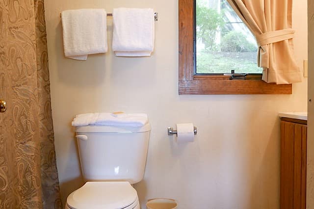 A simple bathroom scene featuring a white toilet with stacked towels, two towels on a rack, a toilet paper dispenser, and a small window.