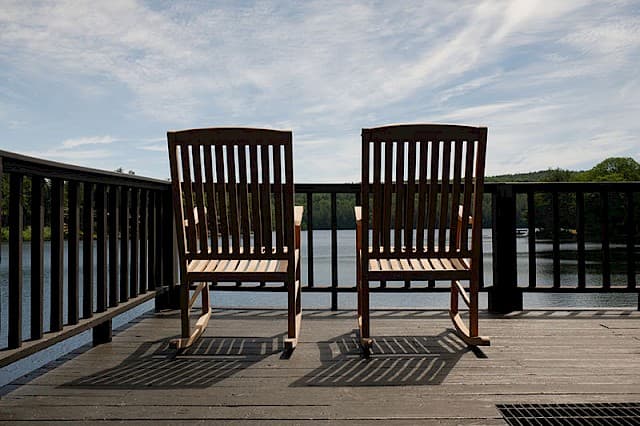 Two slatted wooden rocking chairs sit on a deck overlooking a lake under a bright, cloud-streaked blue sky.