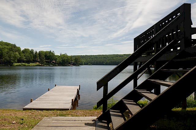 A wooden dock extends into a calm lake, framed by a set of dark stairs and forested banks under a partially cloudy sky.