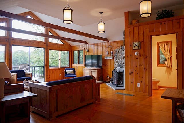 A warm cabin great room with a vaulted ceiling, wood paneling, a stone fireplace, and large windows overlooking a deck.