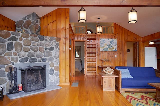 A rustic cabin living room with a vaulted ceiling, hardwood floors, a large stone fireplace, wood paneling, and a loft area.