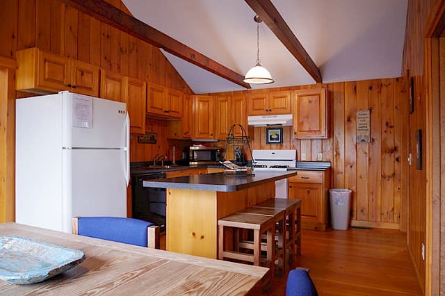A cabin kitchen with wood paneling and a vaulted ceiling with beams, featuring light wood cabinets, a white fridge, and a kitchen island.