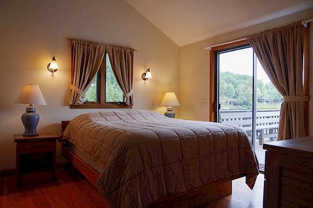 A cozy bedroom with a vaulted ceiling, hardwood floors, a queen bed, and a sliding glass door opening to a deck and natural view.