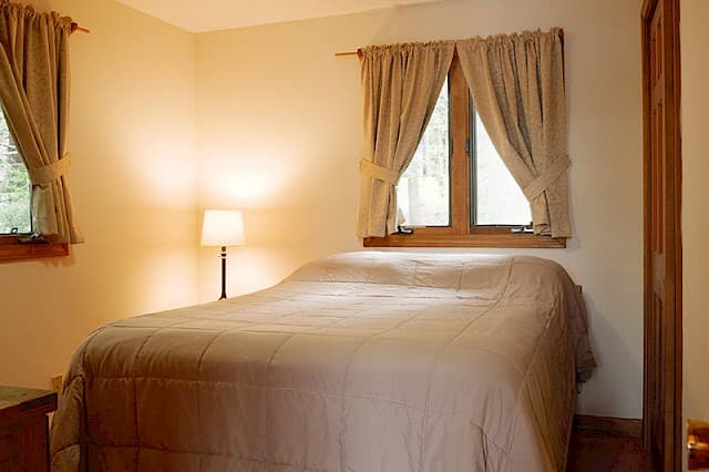 A simple, cozy bedroom with a double bed covered in a neutral comforter, two curtained windows, and a single bedside lamp.