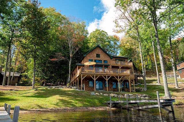 A two-story wooden lake house with a double-decker deck nestled among tall trees with docks extending into the calm water.