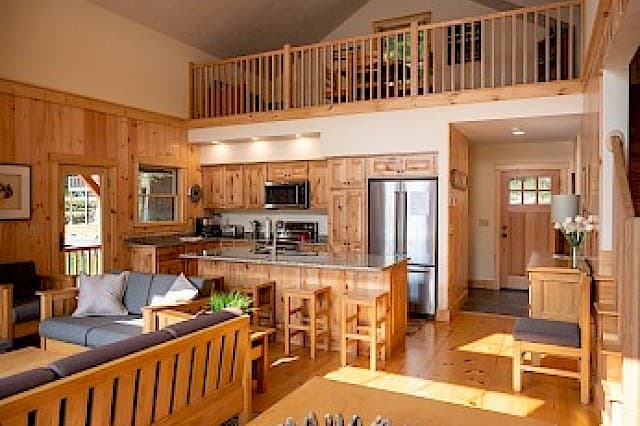 A bright cabin great room with a vaulted ceiling, natural wood paneling, a kitchen island, and a wooden-railed loft overhead.