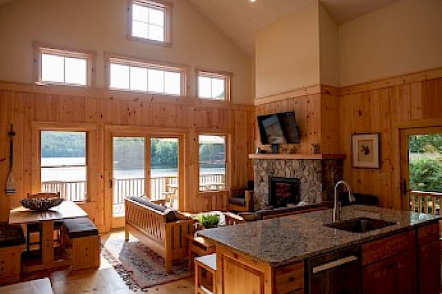 A bright cabin interior with a vaulted ceiling, wood paneling, a stone fireplace, and an island kitchen facing a lake view.